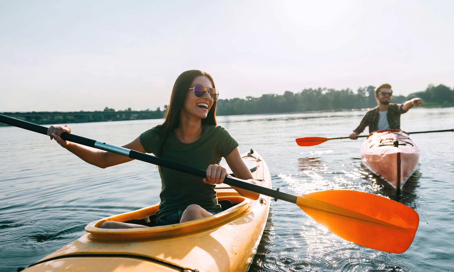 A woman and a man in the foreground, with them are kayaks. They appear to be on a lake or river, enjoying an outdoor activity together.