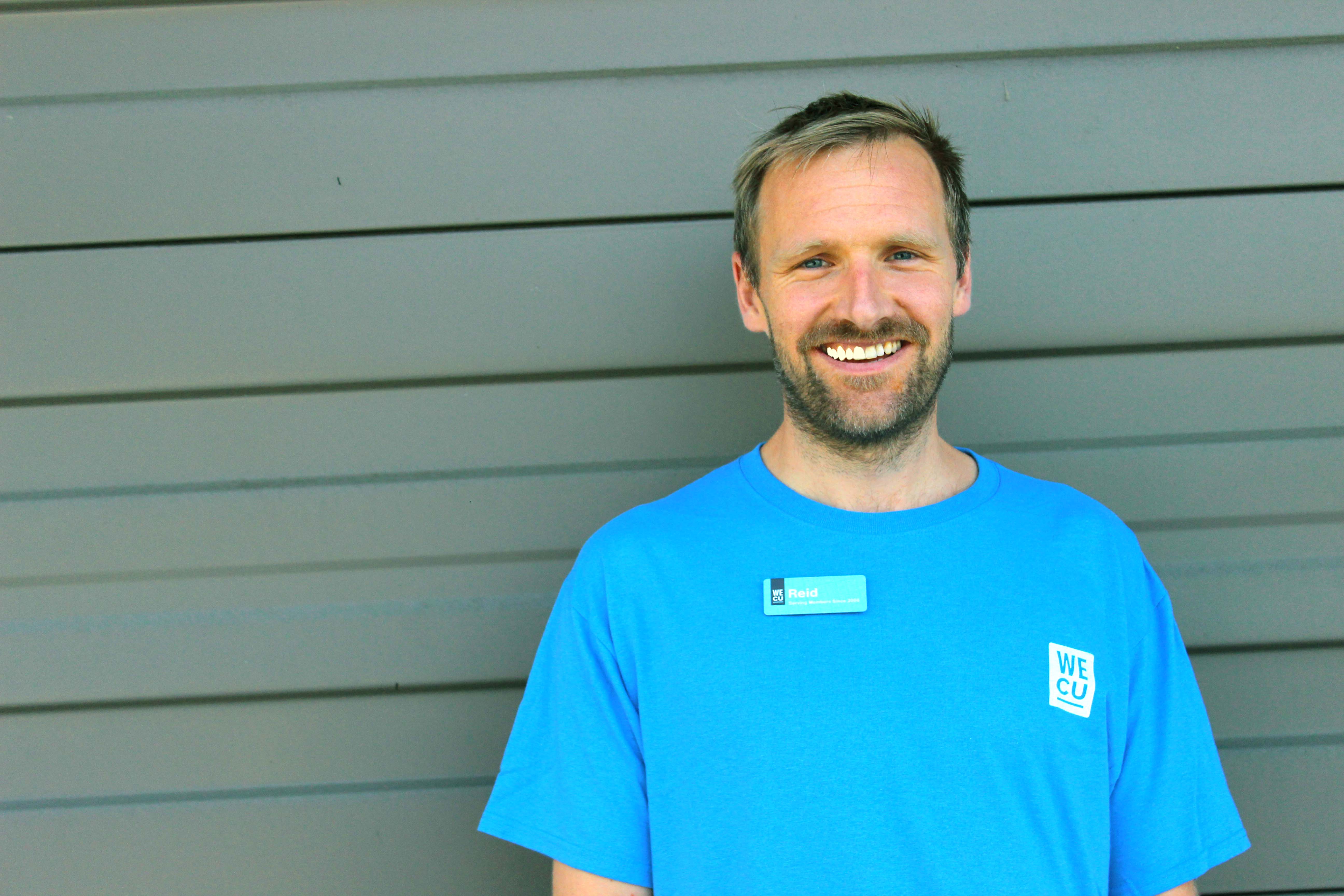 Smiling man wearing a blue t-shirt and posing against a gray wall.