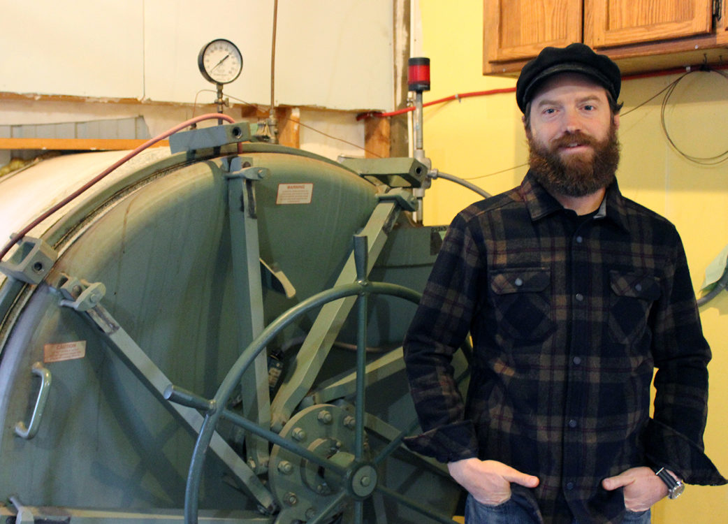 A man in a black plaid shirt stands next to an industrial machine.