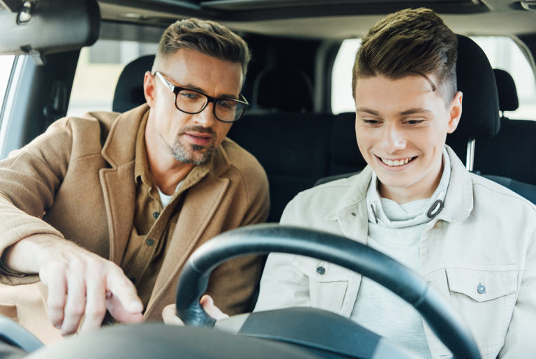 A side view of two men in a car, one man is driving while the other appears to be showing him something.
