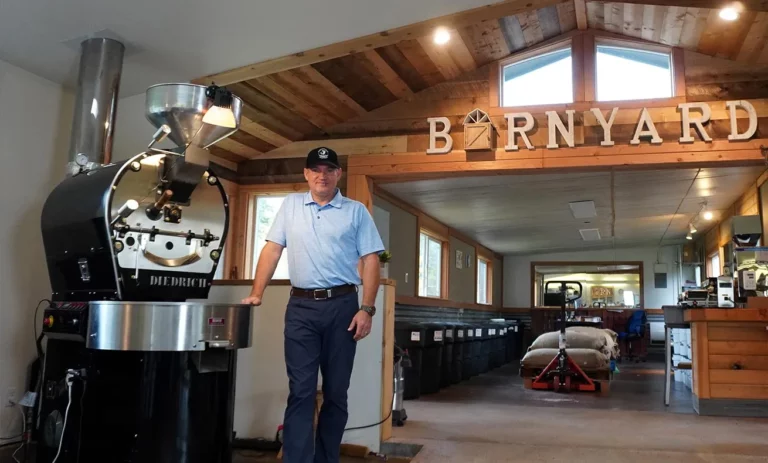 The man is posing next to a coffee machine in front of a sign that says barnyard.