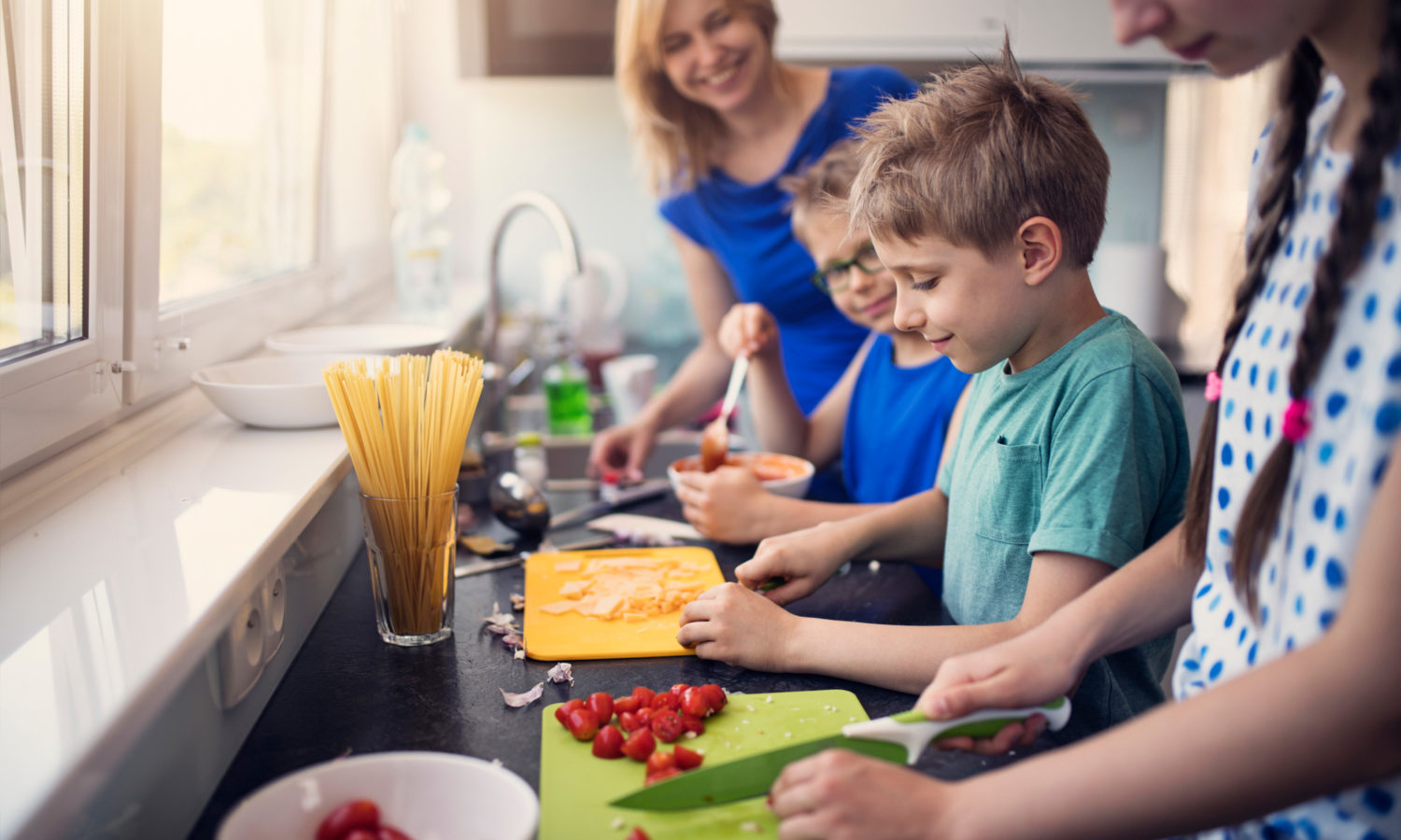 A family of five, including a woman and four children, preparing food together in a kitchen with natural light.