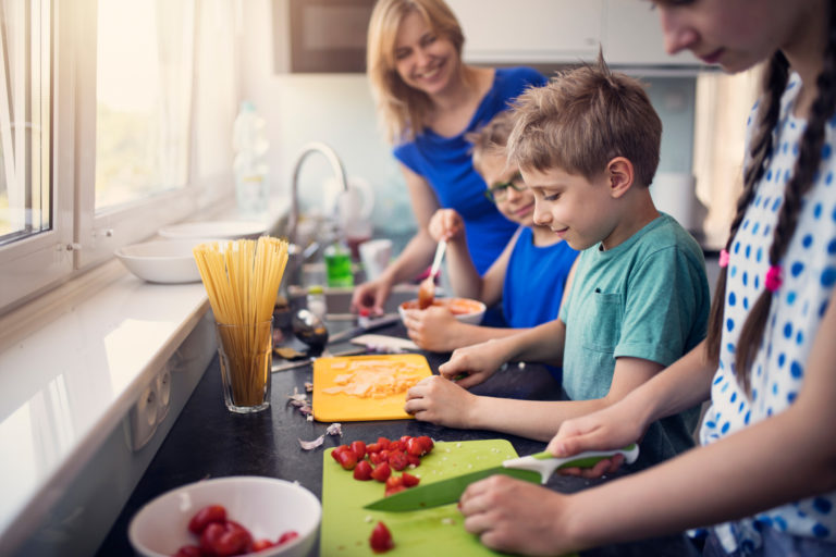 A family of five, including a woman and four children, preparing food together in a kitchen with natural light.