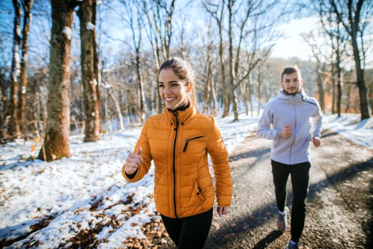 Two people jogging in a wooded area with snow on the ground during daytime.