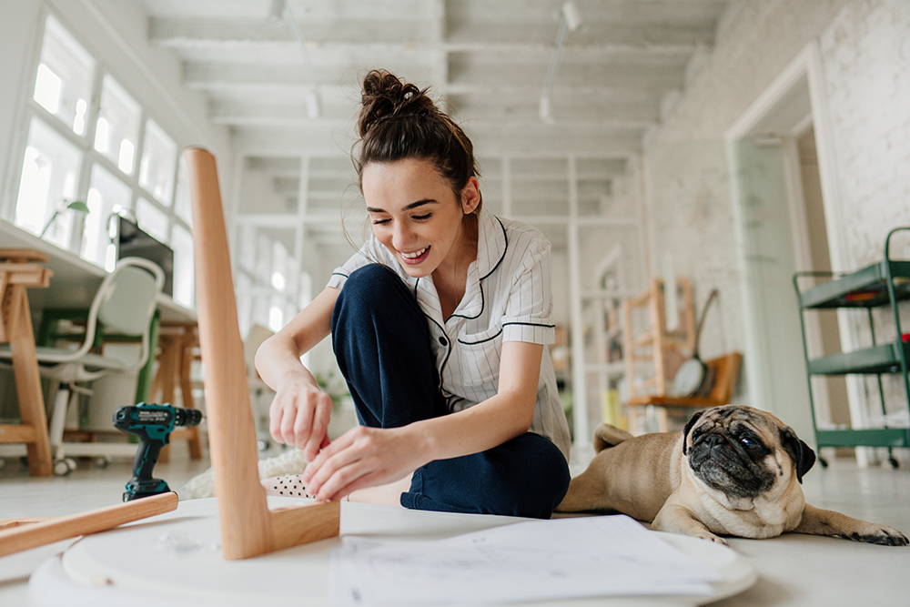 Photo of a smiling young woman assembling furniture, accompanied by her dog in the living room of her apartment.