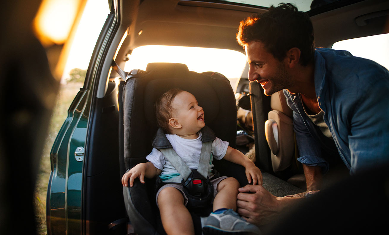 A man smiles at his child who is sitting in a car seat.