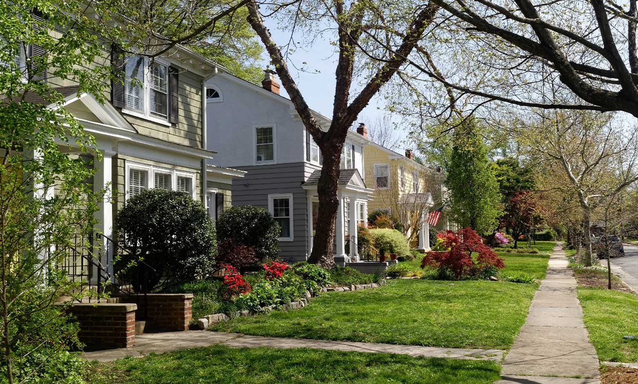 The image depicts a suburban residential street lined with houses, parked cars and a tree.
