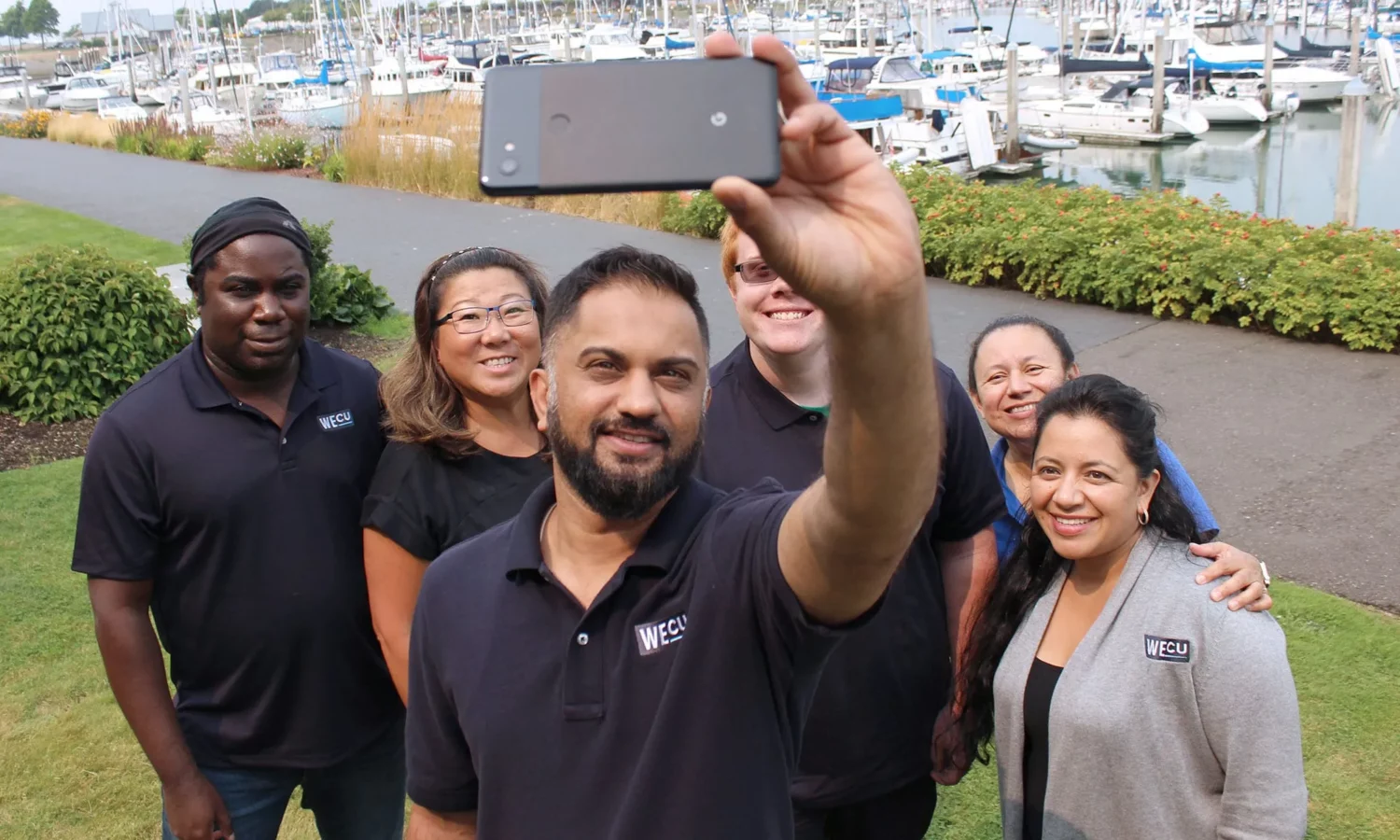 A group of people, including two men and a woman, standing outdoors near water. One man is holding a cellphone while others look on
