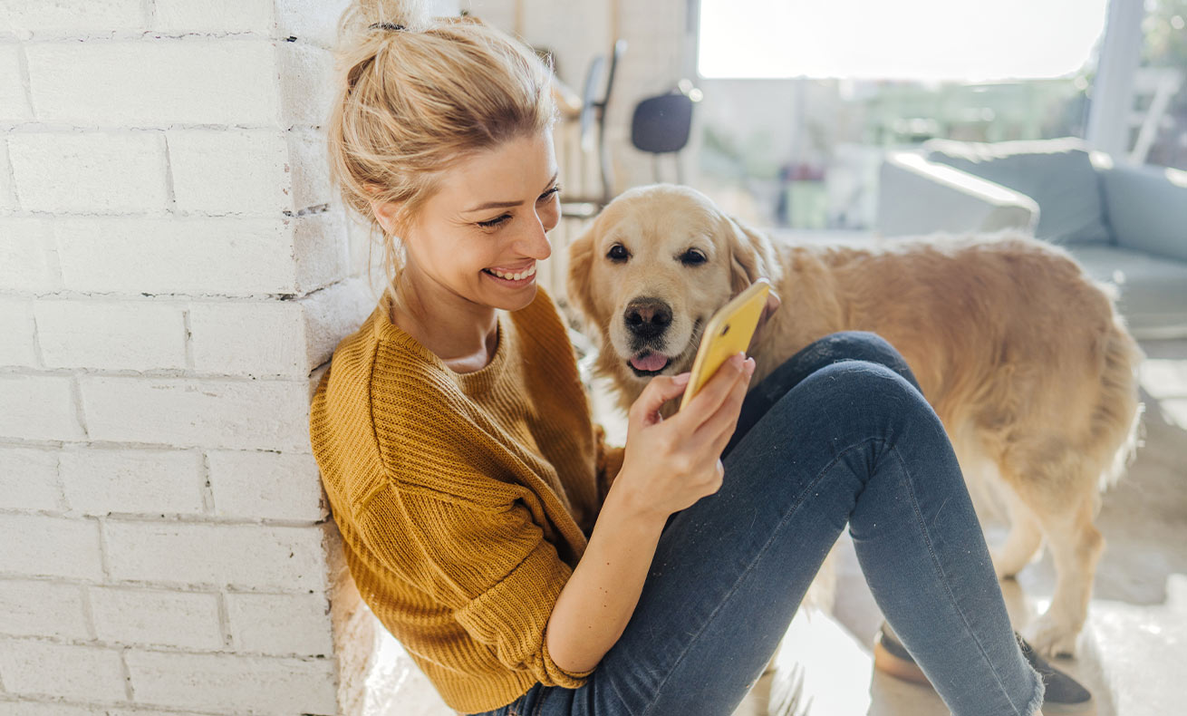 A young woman smiles and looks at her phone while a dog stands nearby.