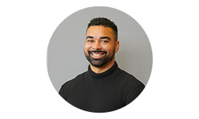 Smiling man with beard and mustache, wearing a black turtleneck, in front of a plain background.