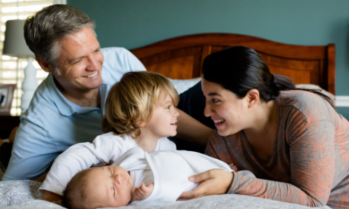 A family photo of a smiling father and mother with their two young children, all in a cozy home environment.