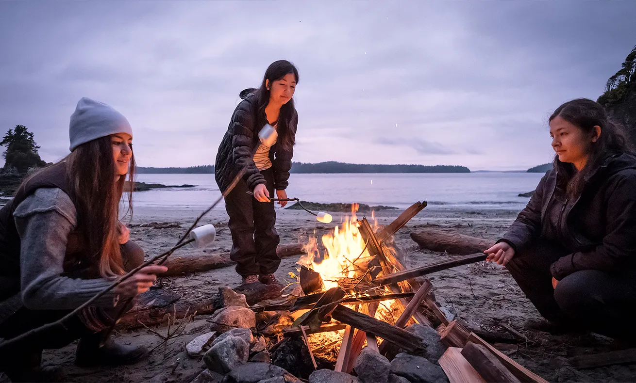 A group of people in winter attire gathered around a fire on the beach.
