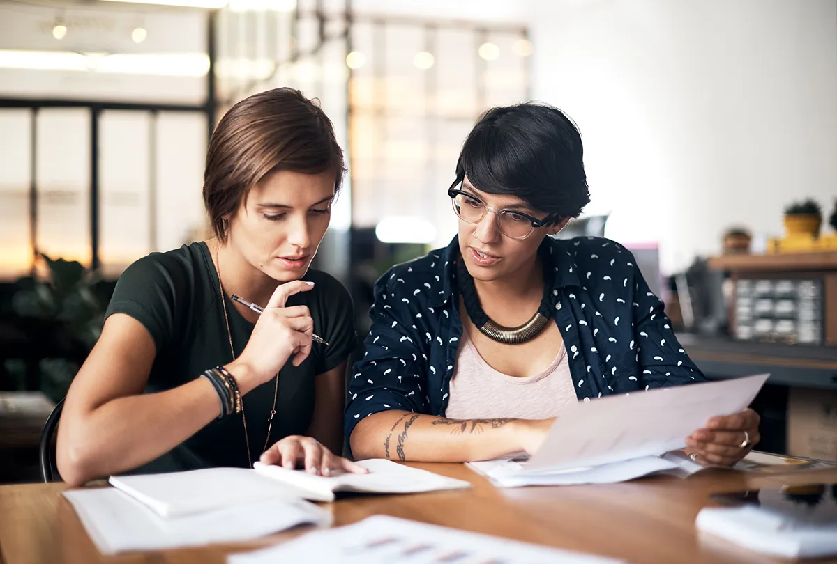 Two people sitting at a table with papers and pens, in an office environment.