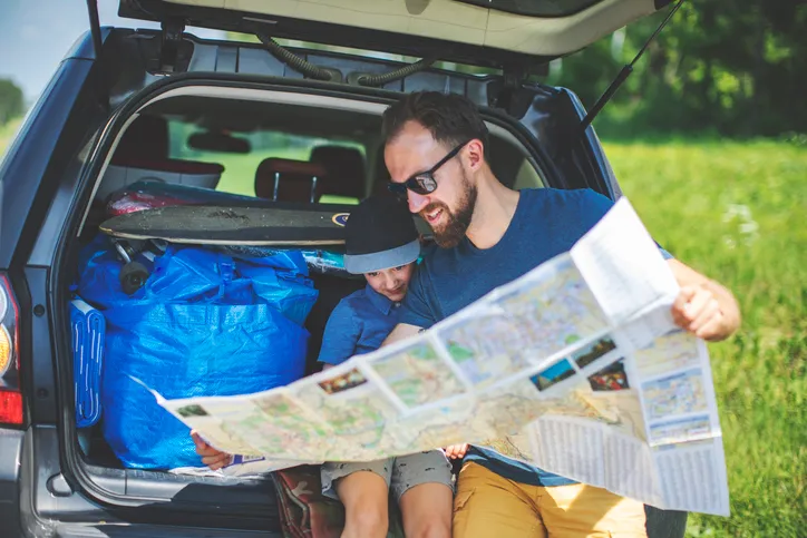 A man with a beard holding an open map in the trunk of a car.