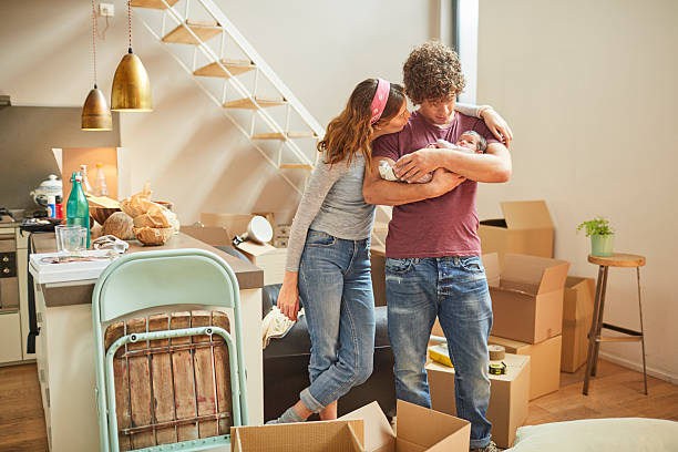 Two people embracing, standing in front of boxes and a staircase in a room with white walls.