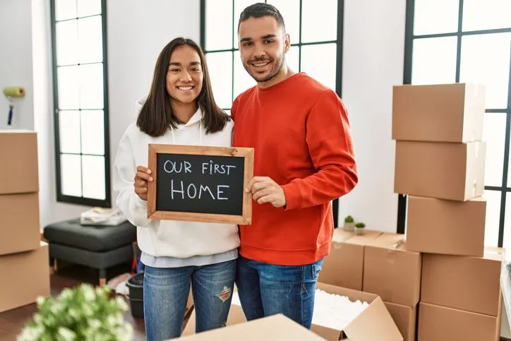 Couple in front of cardboard boxes holding sign saying Our first home.