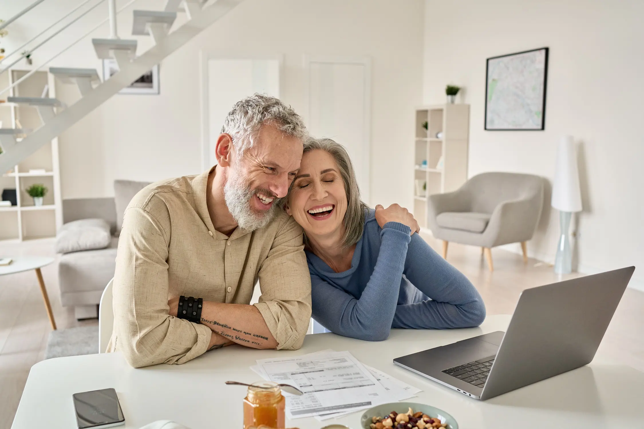 A man and woman sitting at a table in a living room, smiling and looking at the camera, with a laptop between them.