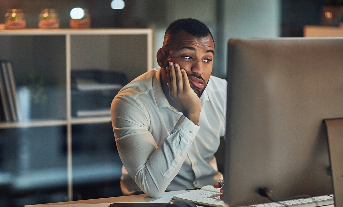 A man sitting at a desk, looking down and holding his head in both hands.