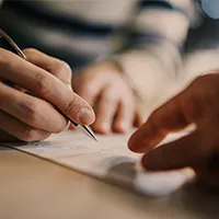 A pair of hands signs a document as another points where to sign for their business loan.